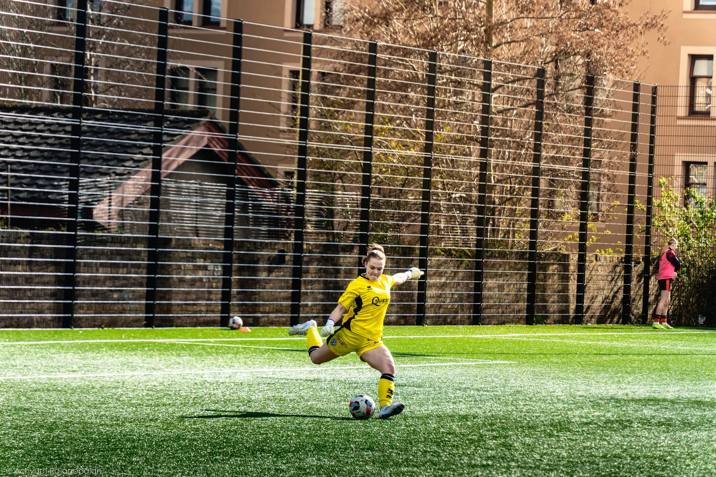 A female soccer goalkeeper in a yellow uniform is in action on a green artificial turf field, making a save as she kicks the ball away. In the background, a playground, trees, and apartment buildings are visible, with a few other individuals watching