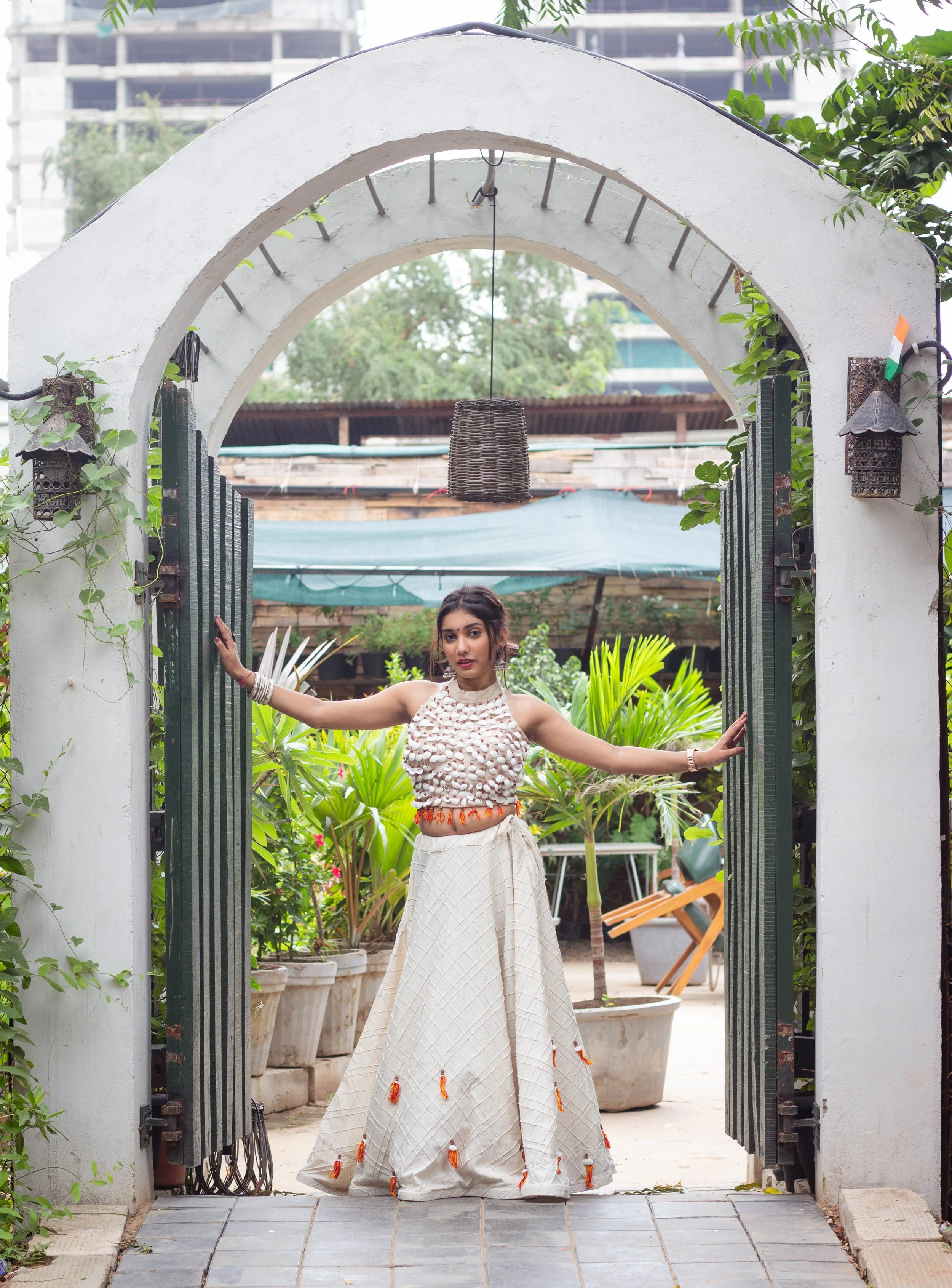 A woman dressed in traditional Indian attire standing in an arched garden gate, surrounded by greenery and outdoor furniture. She is holding open the gate with her arms extended, wearing a beige and orange outfit with jewelry, in a setting with potted plants and a wooden structure in the background.