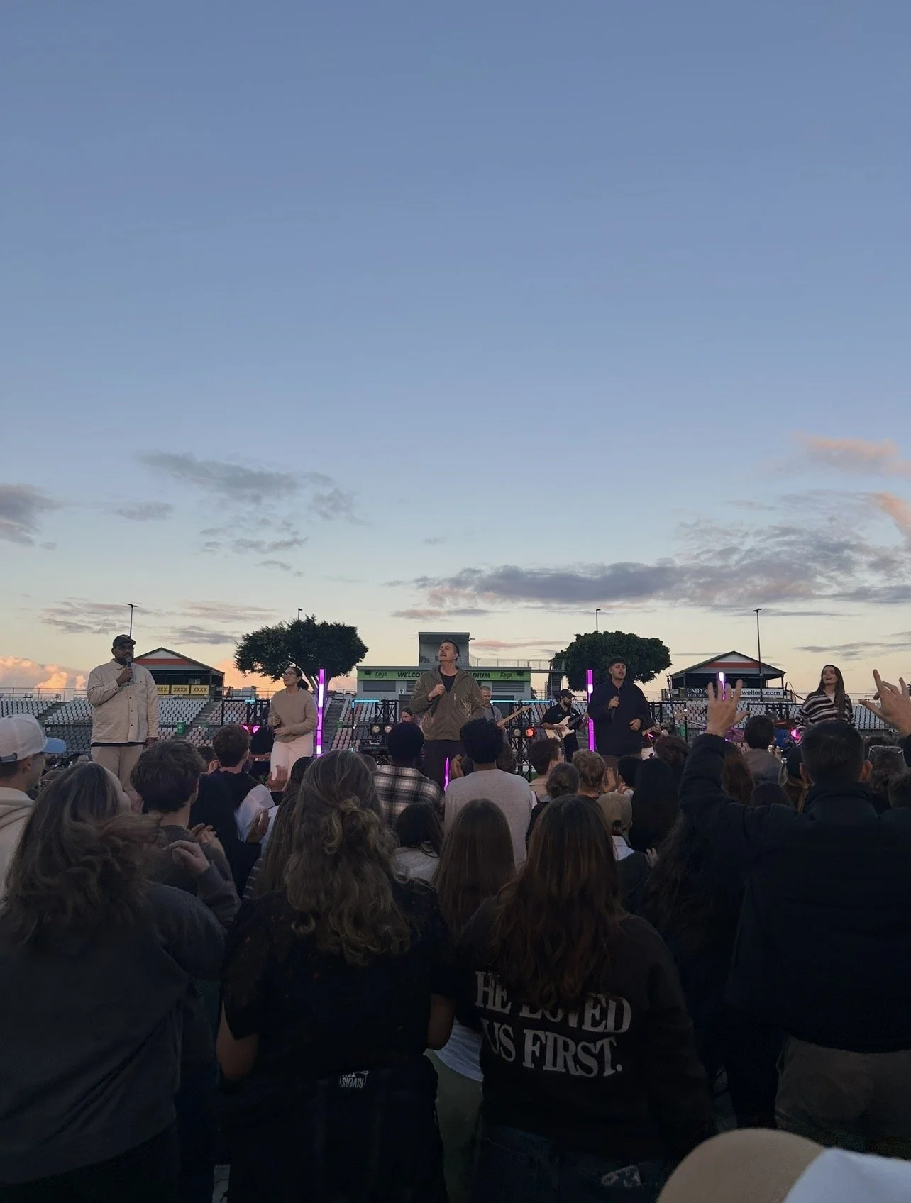 A group of people performing on stage at an outdoor concert during sunset, with an audience watching.
