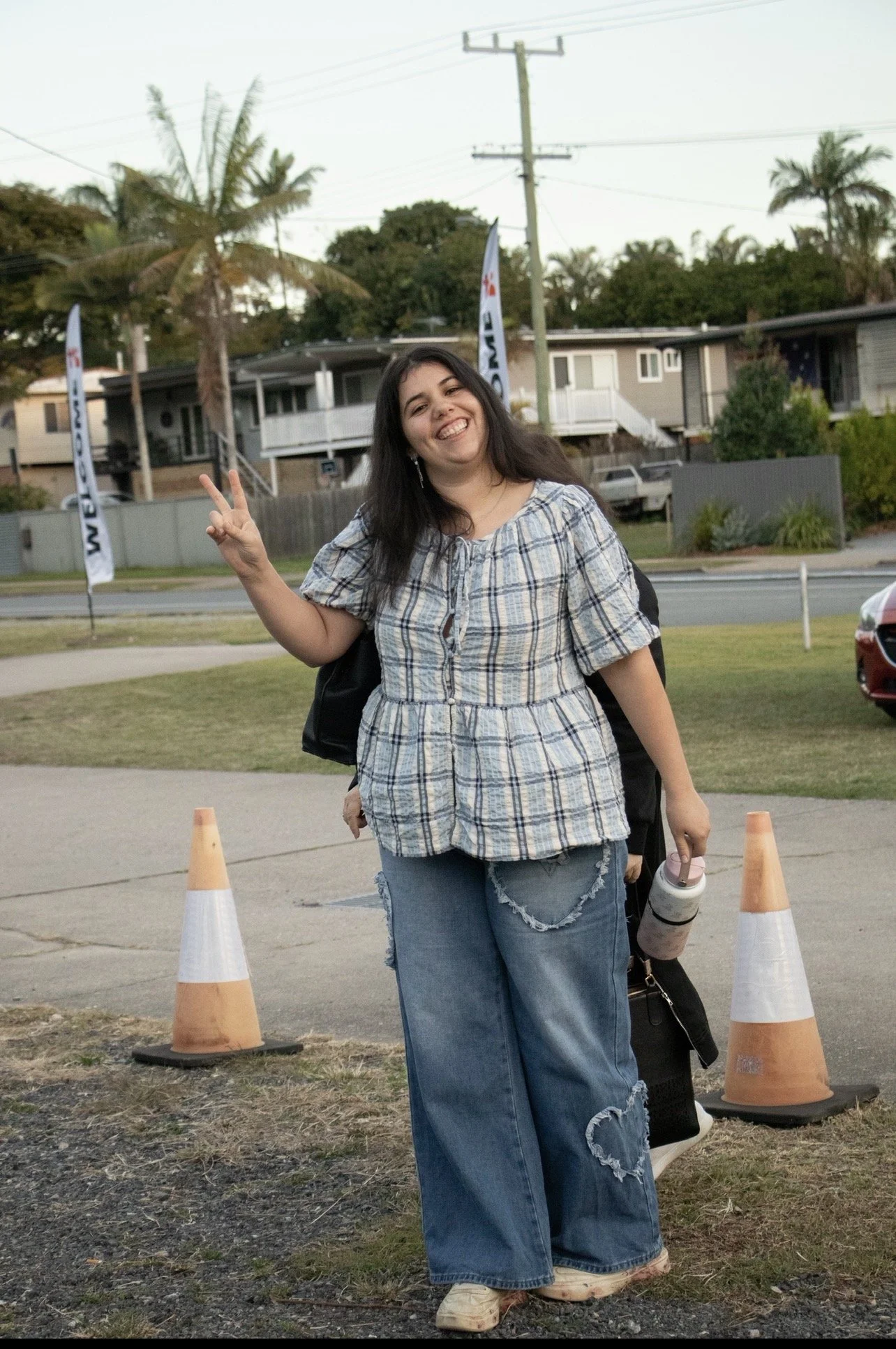 A young woman standing outdoors on a sidewalk, smiling and making a peace sign with her right hand. She is wearing a plaid shirt and ripped jeans, with a backpack on her back, and holding a water bottle. There are orange traffic cones in front of her