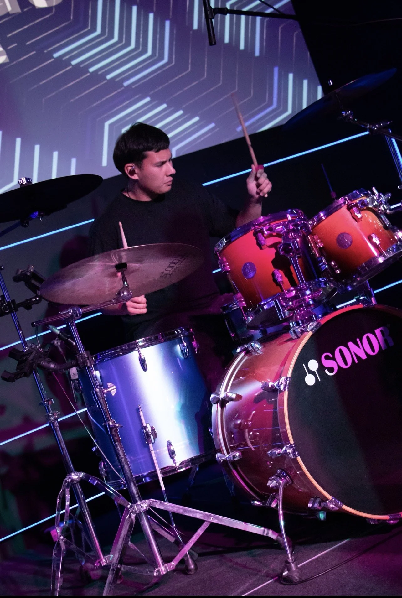 A man playing a red drum set on stage during a live performance, with a digital geometric background projection.