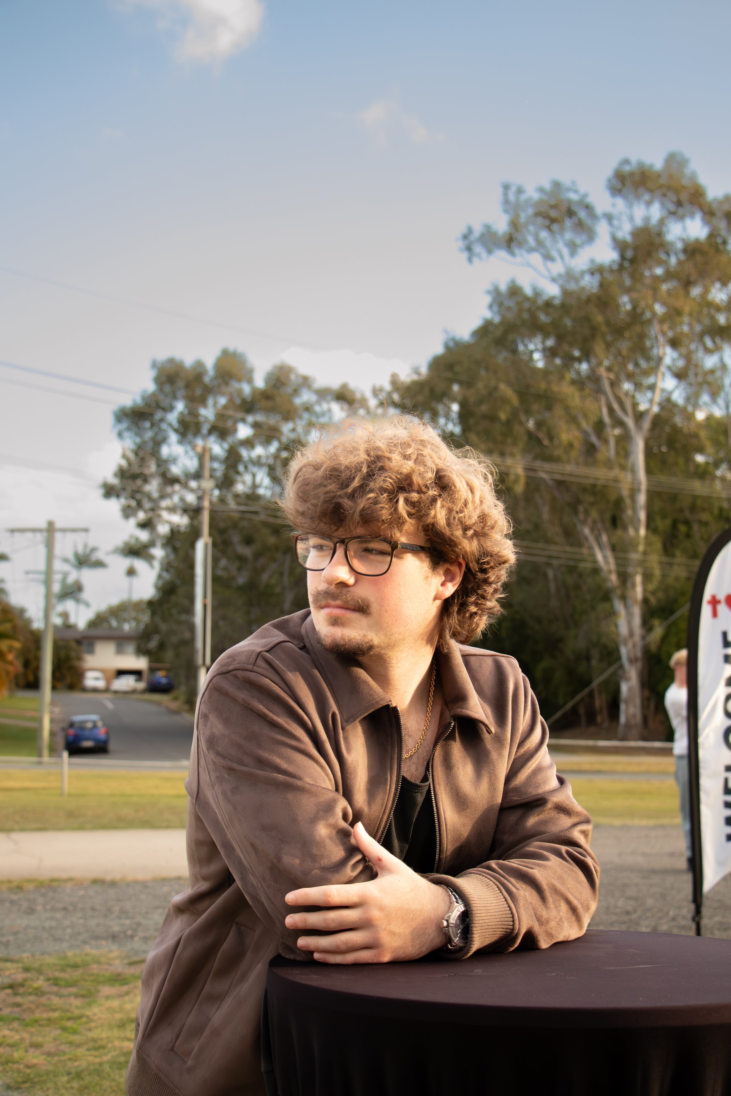 A young man with curly hair and glasses wearing a brown jacket, sitting outdoors with trees and a parking lot in the background.