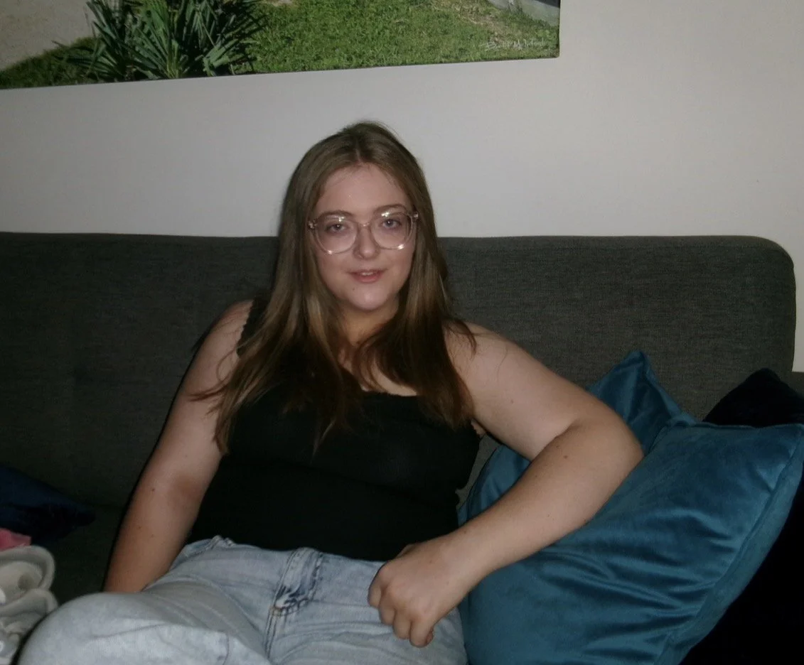 A young woman with long brown hair and glasses sitting on a gray couch with blue pillows, wearing a black top and light jeans, in a room with a white wall and a framed picture behind her.