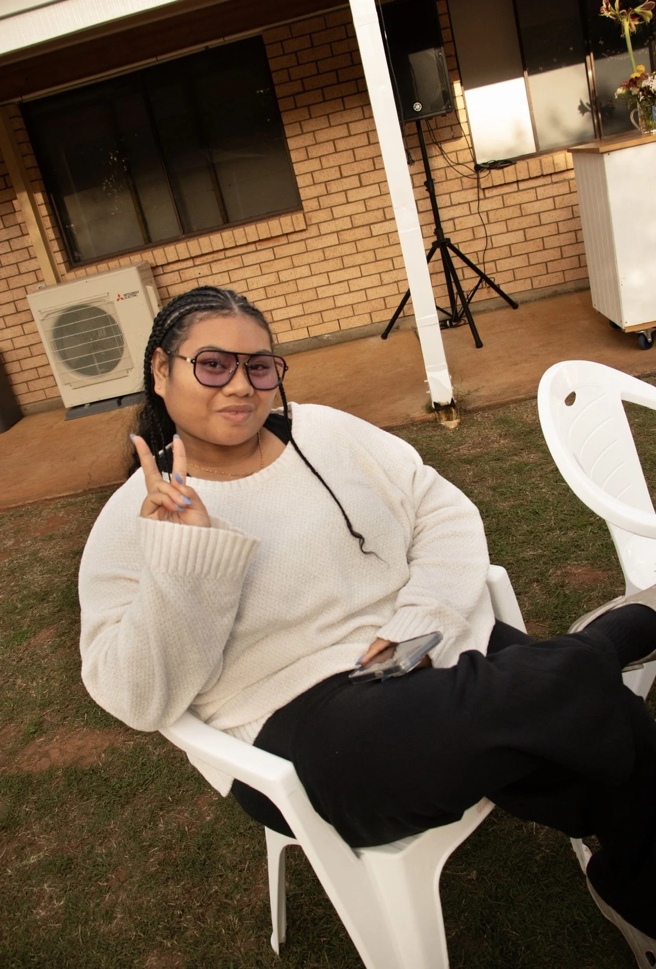 A woman with braided hair wearing sunglasses, a cream-colored sweater, and black pants, sitting outdoors on a white plastic chair, showing a peace sign with her fingers, holding a phone in her lap, in front of a brick house with a window and an air c