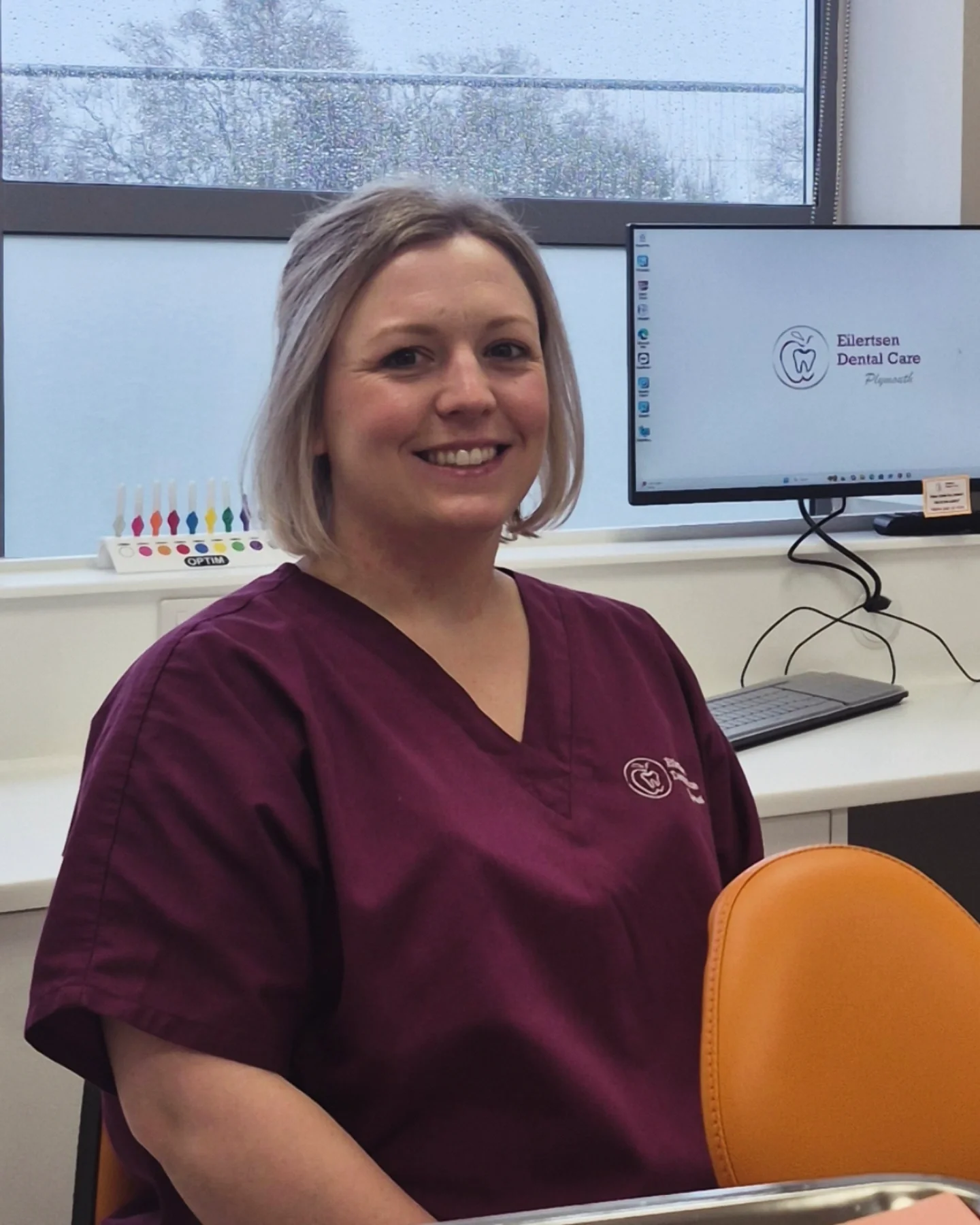 Gemma is sat behind an orange dental patient chair, in maroon scrubs. she is smiling.