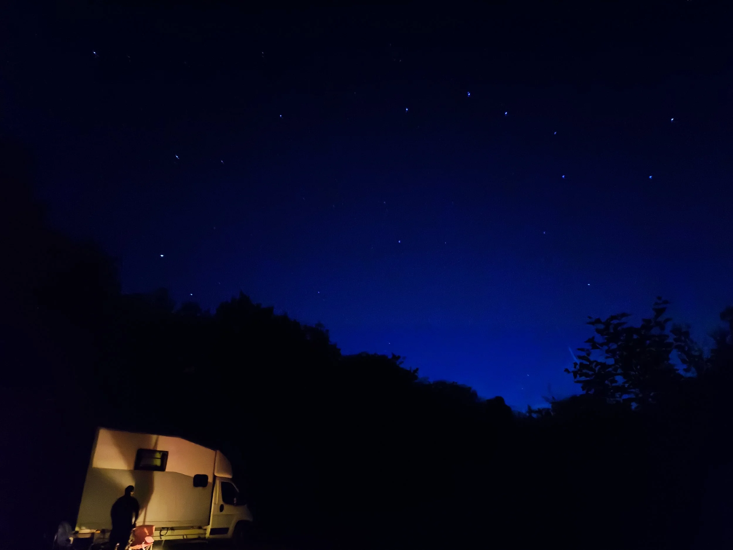campervan with nightskyline of trees, stars in sky, fire in front of the van with a person casting shadows
