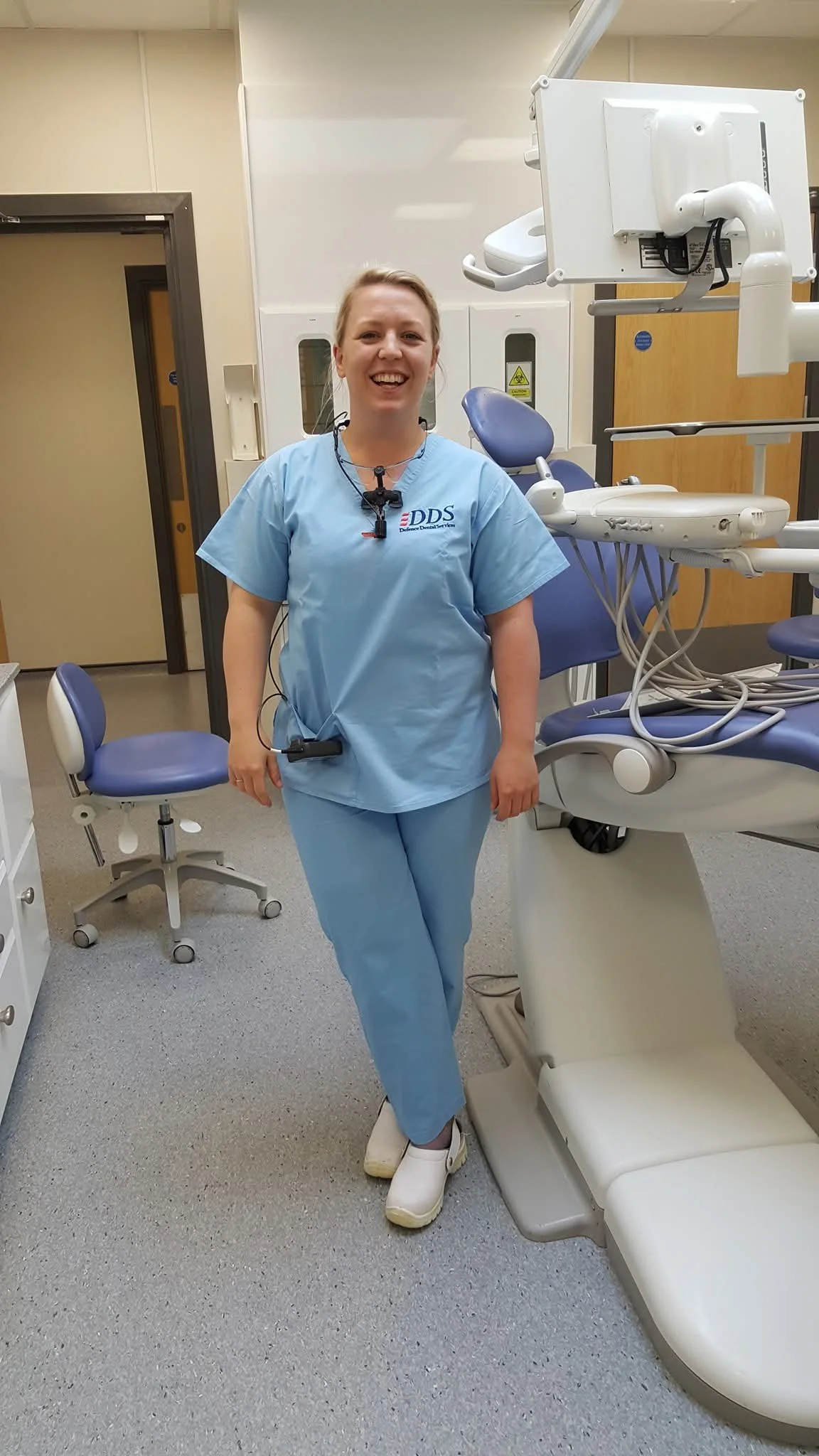 photo of Gemma standing in a dental surgery with blue DDS (defence dental service) scrubs. Gemma is smiling.