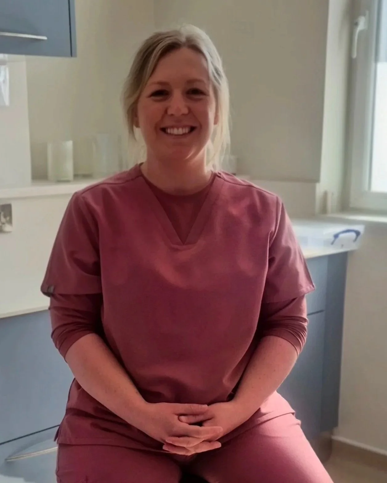 Photo of Gemma in a dental surgery. A woman with blonde hair smiling, wearing pink medical scrubs, sitting in a medical or clinical setting with a window and cabinets in the background.