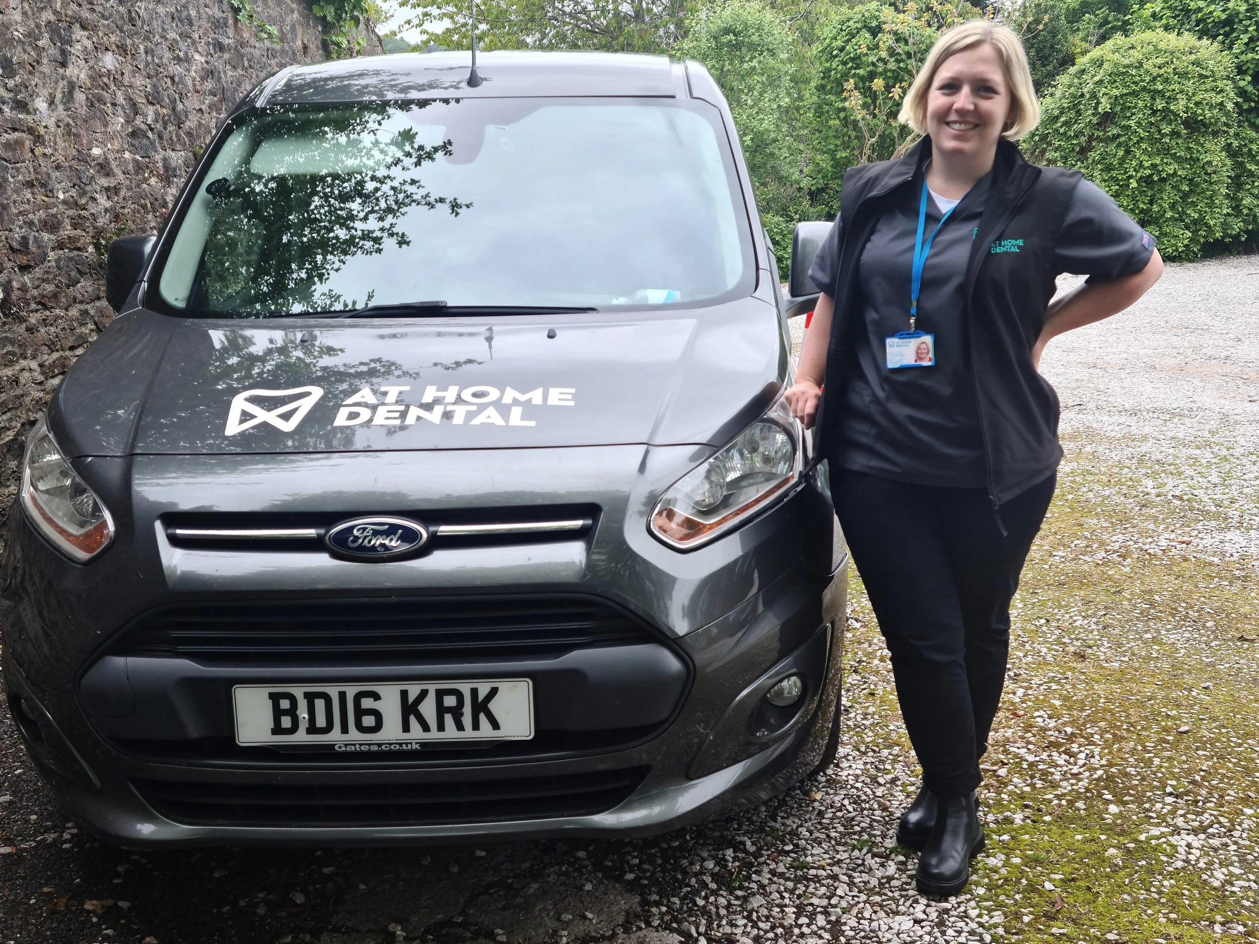 Gemma standing in a car park next to a van in scrub top, gilet and lanyard with black trousers and boots. The gilet and the van both have  "At Home Dental" branding