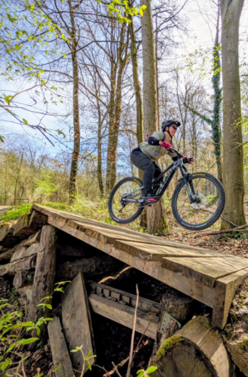 Gemma on a mountain bike, slightly off the ground, coming off a wooden jump, in full mountain bike gear