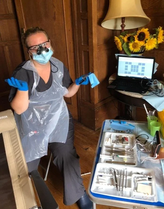 A photo of Gemma set up for dentistry in someone's home. wearing a face mask, loupes, gloves, and a plastic apron, standing in a room with wooden walls next to a table with dental tools and a computer monitor displaying a calendar.