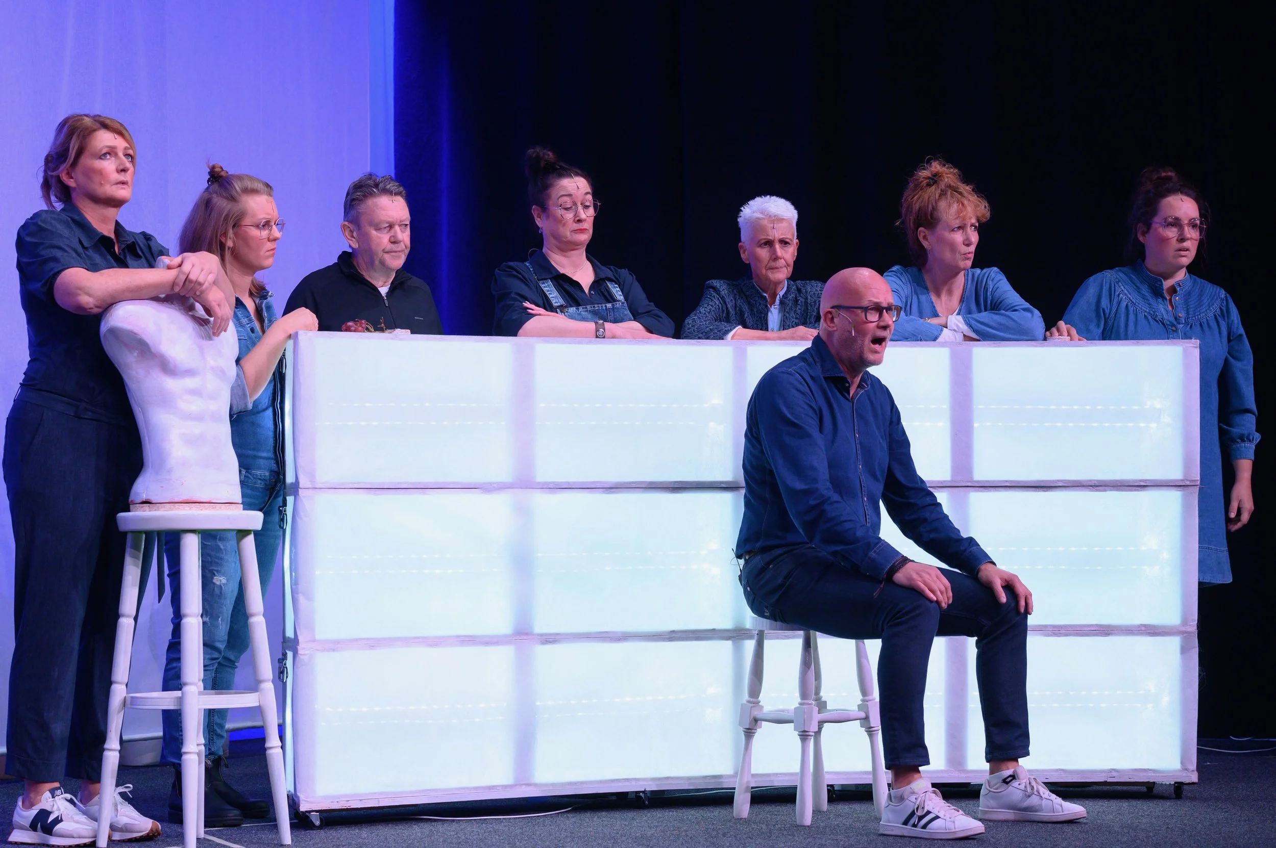 Een groep mensen staat en zit op een toneel, met uitdrukkingen van bezorgdheid of verwachting, mogelijk tijdens een toneelstuk of scène.