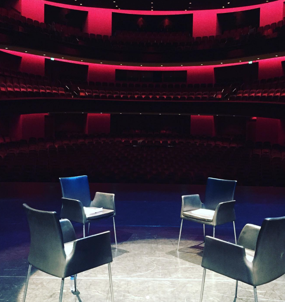 Vier lege zwarte stoelen op een donkere theater- of congreszaal met rode verlichte boksen en meerdere kijkersrekken.