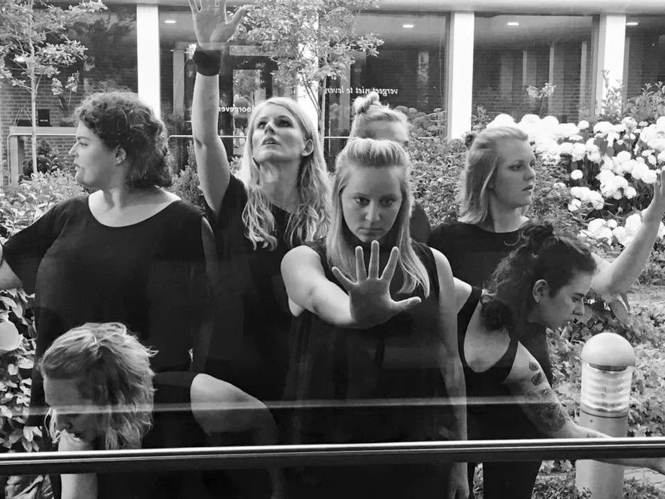 Vijf vrouwen die achter een glazen raam poseren, met verschillende uitdrukkingen en handgebaren, in een tuin met bloemen en planten buiten.