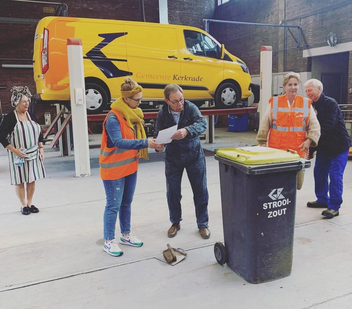 Vrouw met een oranje veiligheidsvest en een gele sjaal kijkt naar een man die een papiertje vasthoudt, terwijl andere mensen in veiligheidskleding en een persoon in een feesthoed op de achtergrond bezig zijn in een loods met een grote gele vrachtwagen.