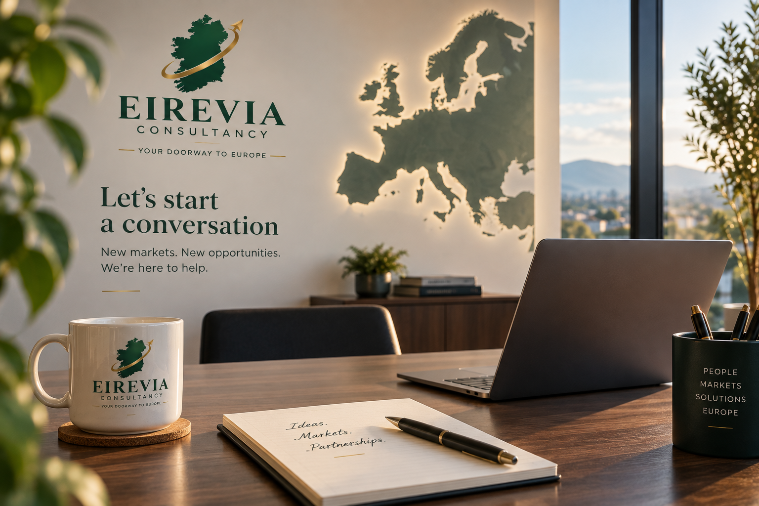 Office workspace with Eirevia Consultancy branding, a laptop, mug, notepad, pens, and a window view of a city and mountains, with a European map on the wall.