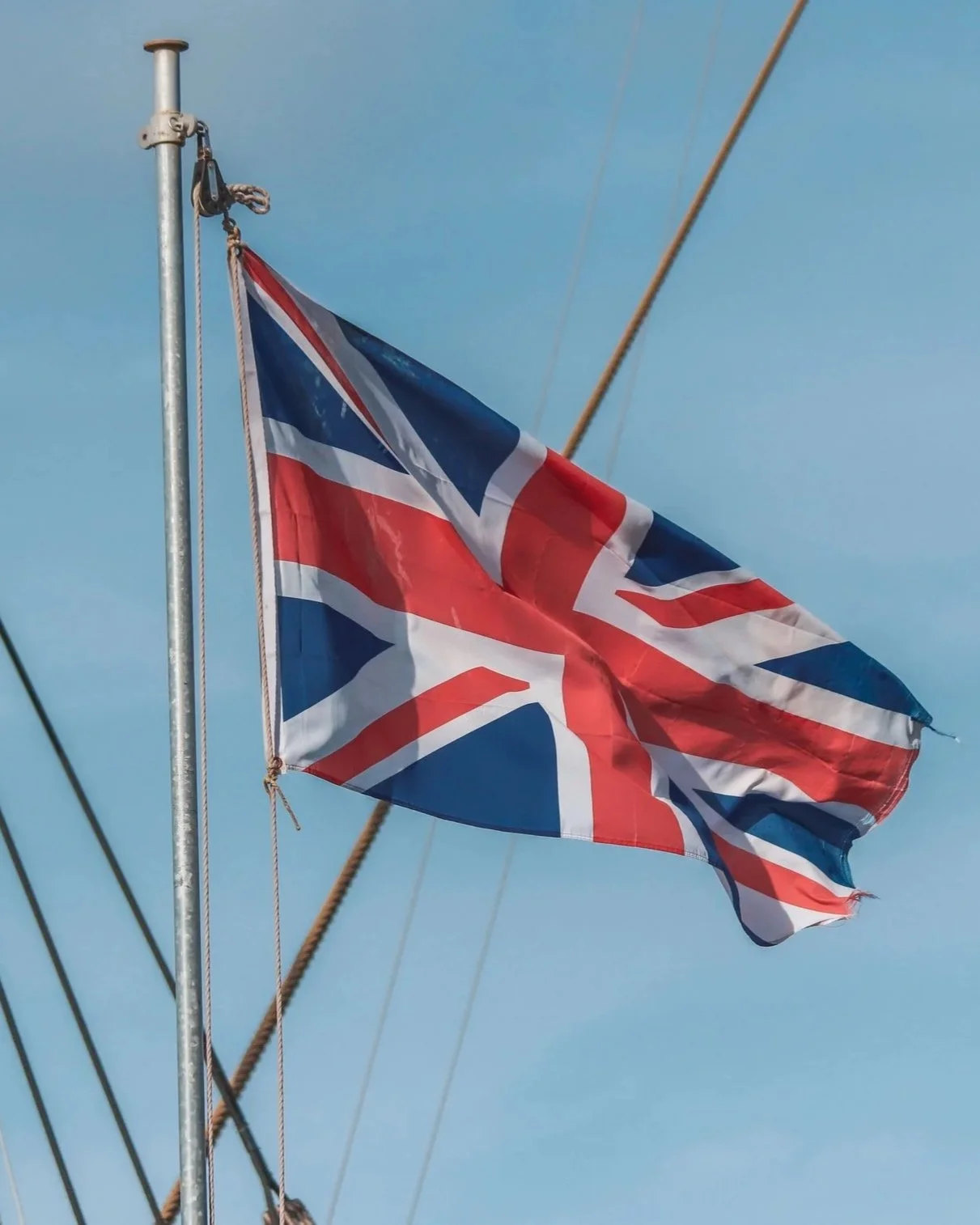 A Union Jack flag flying outdoors against a blue sky, attached to a metal pole with ropes and wires visible in the background.