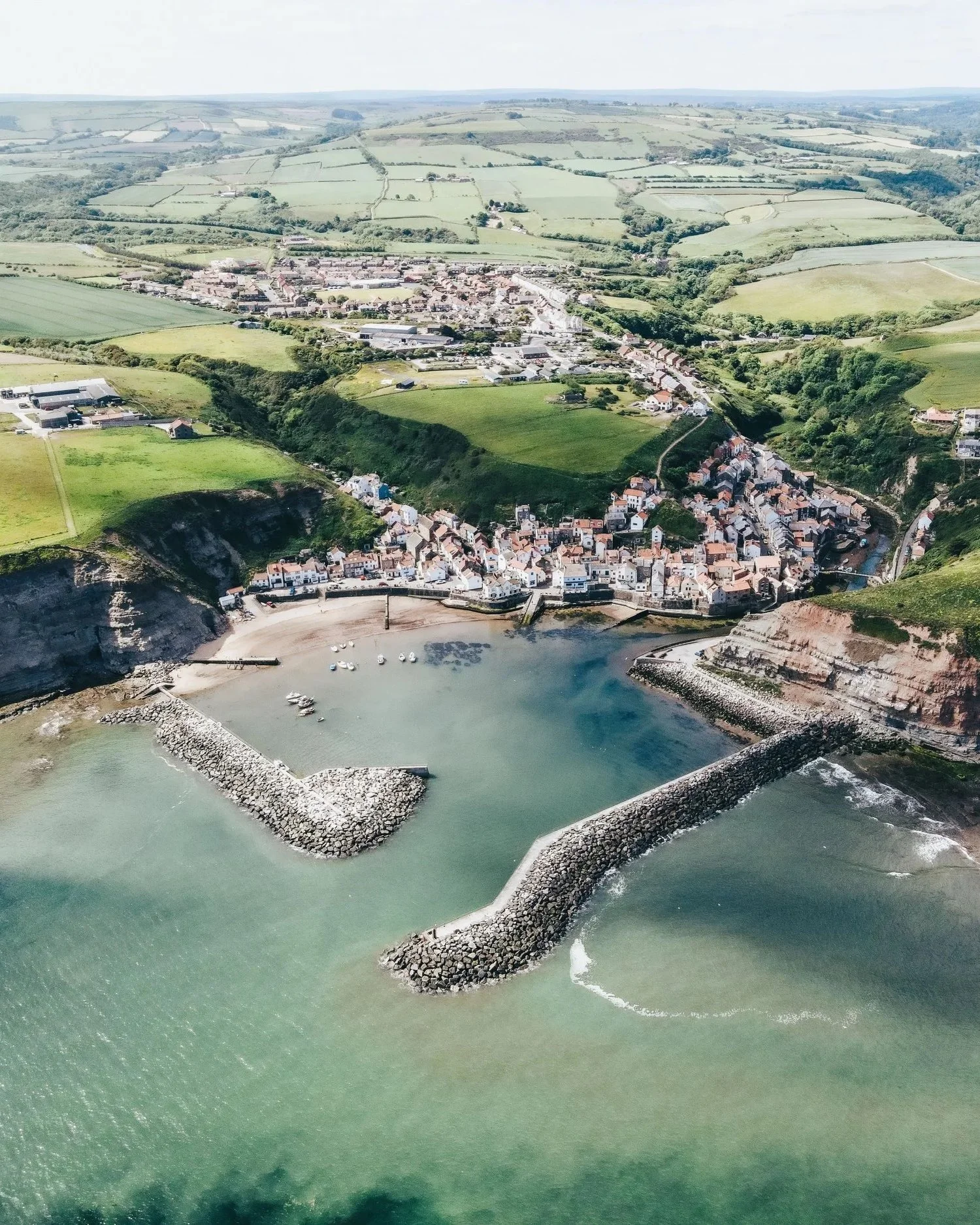Aerial view of a coastal town with a harbor protected by stone breakwaters, houses on the shoreline, and agricultural fields and rolling hills in the background.