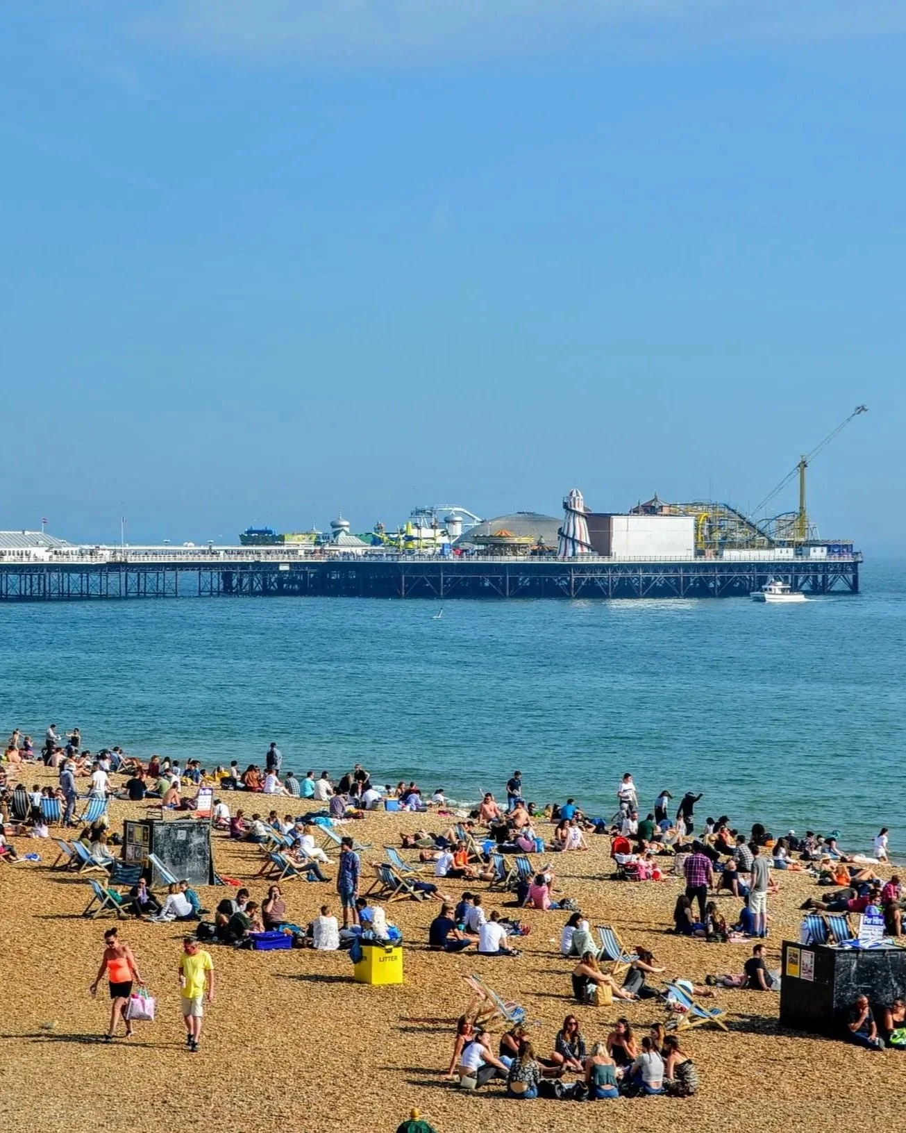 People relaxing and sunbathing on a crowded beach in Brighton with lounge chairs and umbrellas, near a pier extending into the ocean, with amusement park rides visible on the pier and a boat in the water.