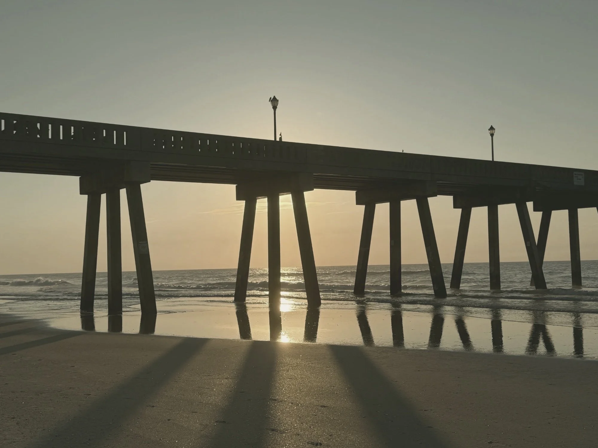 Sun rising through the pilings of an ocean pier, reflected on wet sand