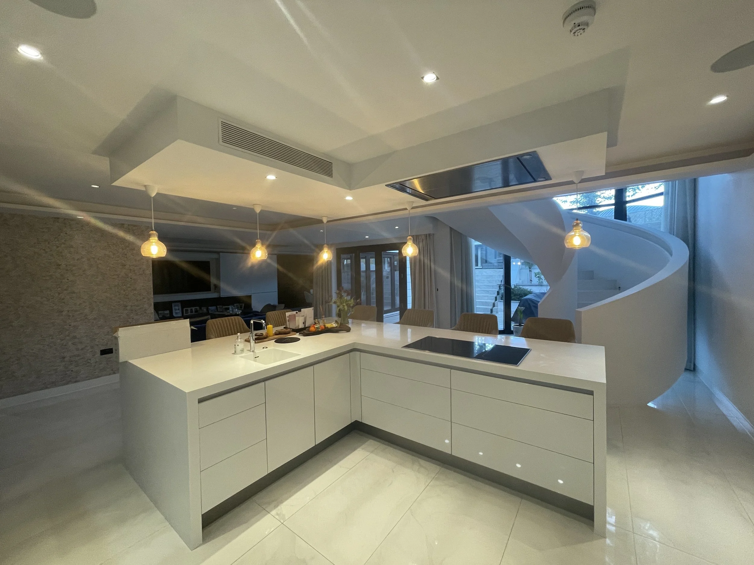 Modern kitchen with white island, induction cooktop, pendant lights, spiral staircase, and large windows in the background.