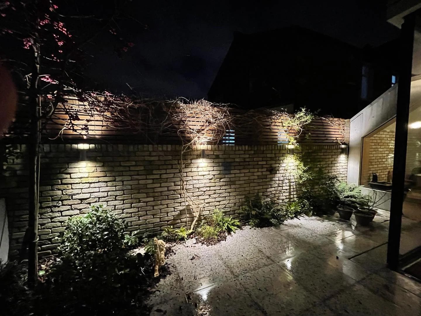 Night view of a backyard patio with a brick wall, garden plants, and outdoor lighting illuminating the area.