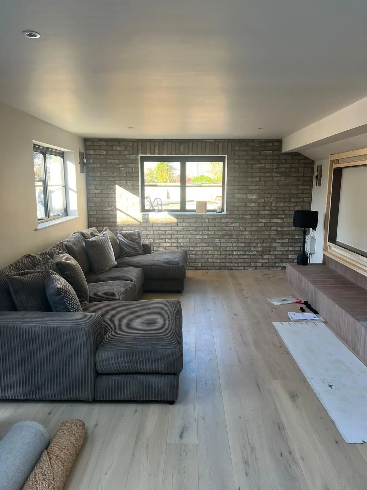 Modern living room with gray sectional sofa, exposed brick accent wall, windows, light wood flooring, and built-in TV console
