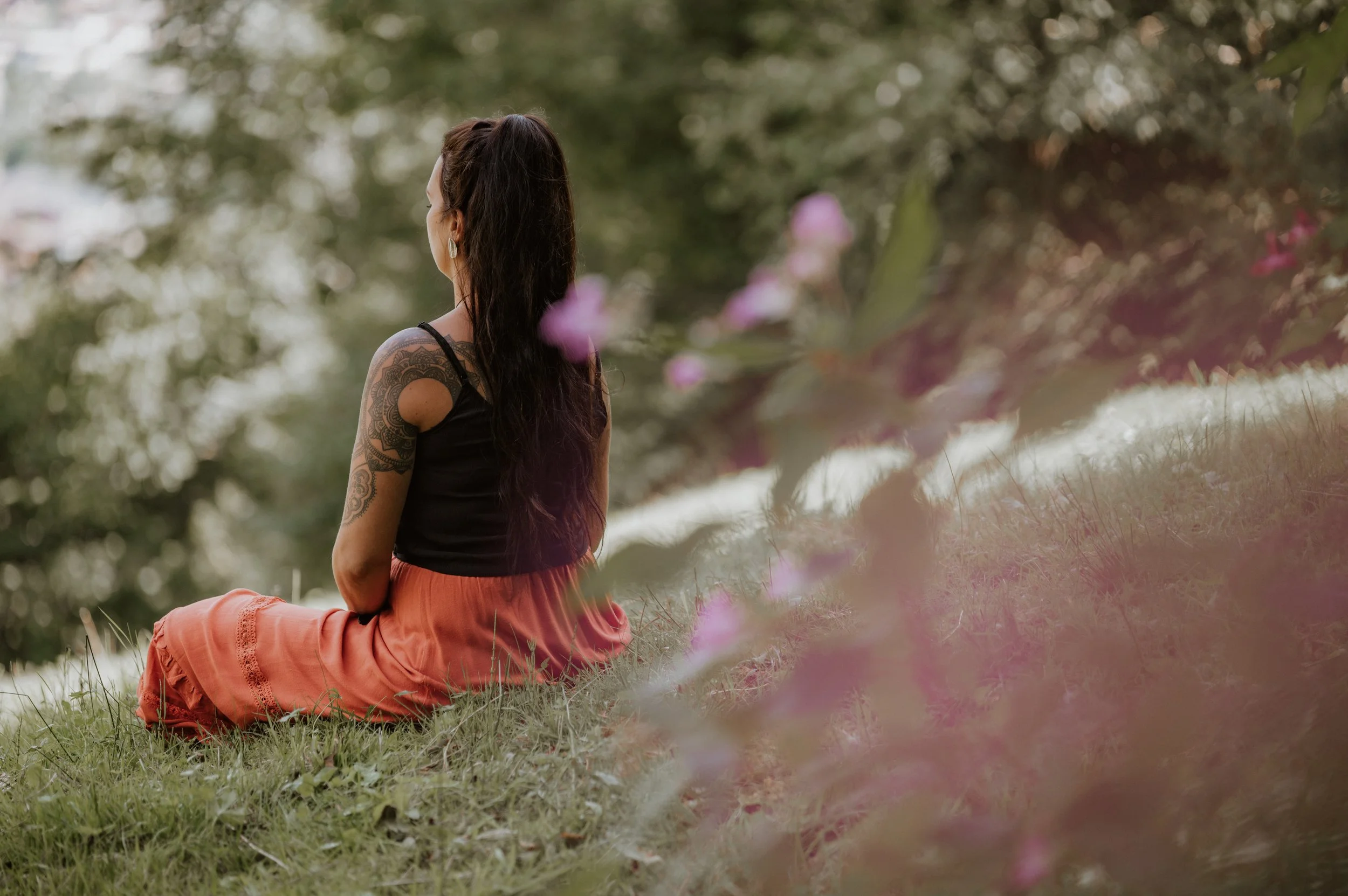 Eine Frau mit langen dunklen Haaren und Tattoos am Arm sitzt im Gras und schaut in die Ferne in einer natürlichen, grünen Umgebung mit Bäumen und Blumen.