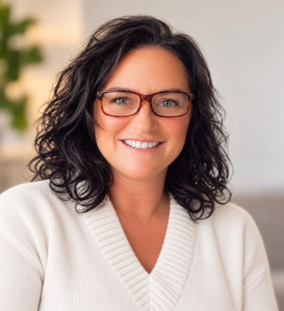 Smiling woman with curly dark hair, wearing glasses and a white sweater, indoors with a blurred background.
