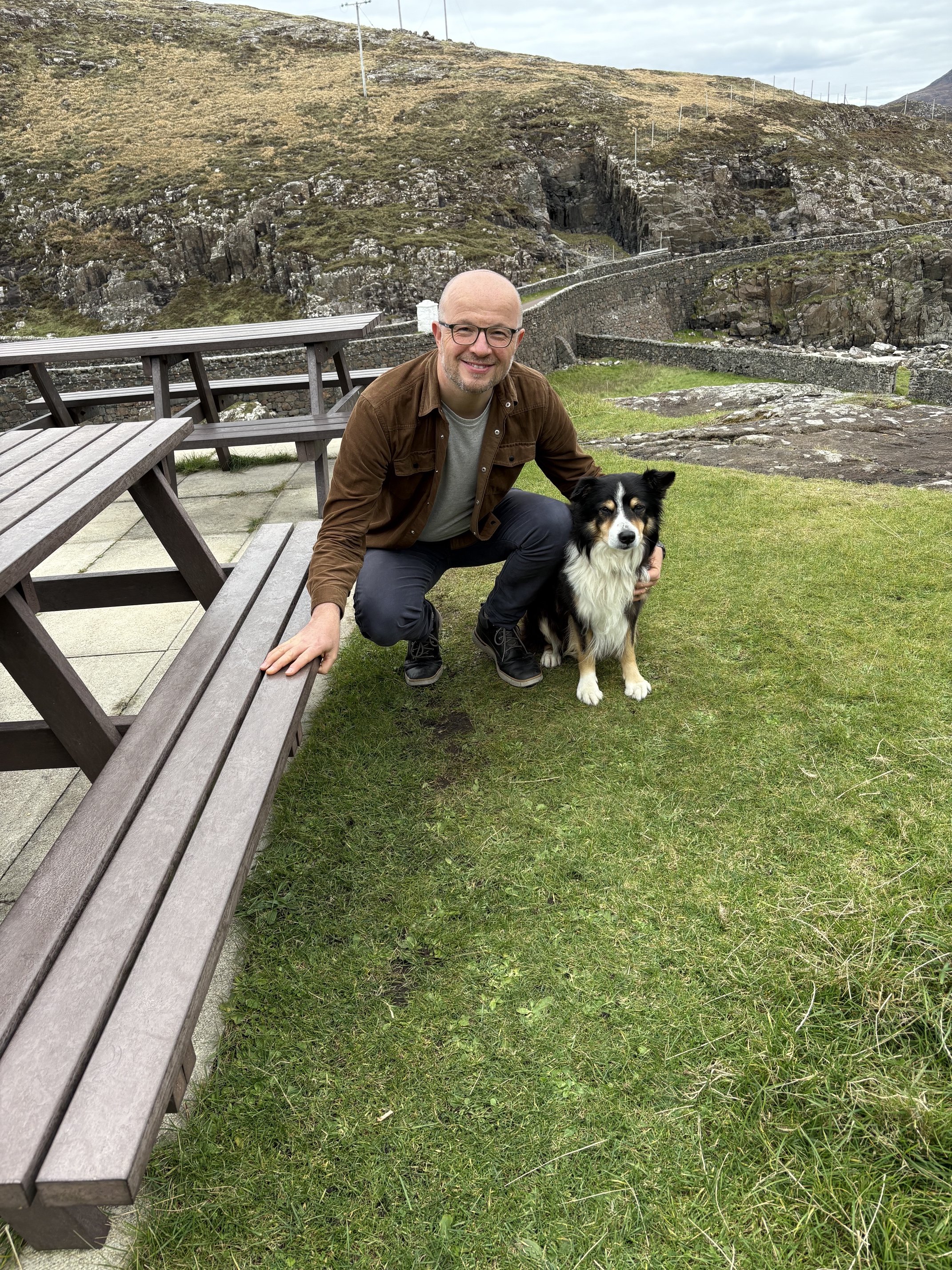 A man wearing glasses, a brown jacket, and dark pants crouches next to a black and white border collie dog on green grass. They are outdoors with a rocky, hilly landscape and a stone fence in the background.