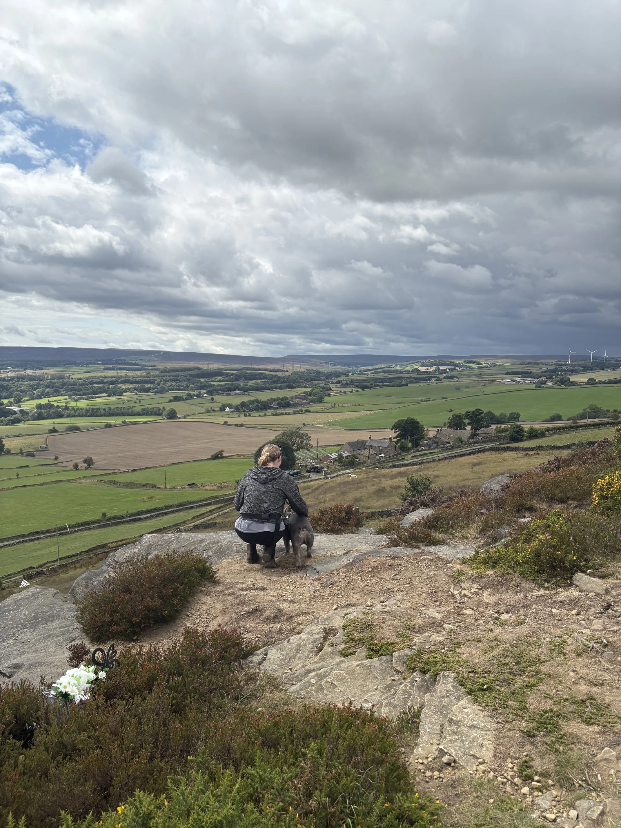 A woman in gray clothing walking with her dog on a rocky trail in a green rural landscape under a cloudy sky with wind turbines in the distance.