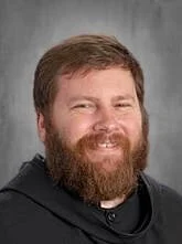 A man with a beard and short hair, smiling, wearing a dark hoodie, standing against a gray background.