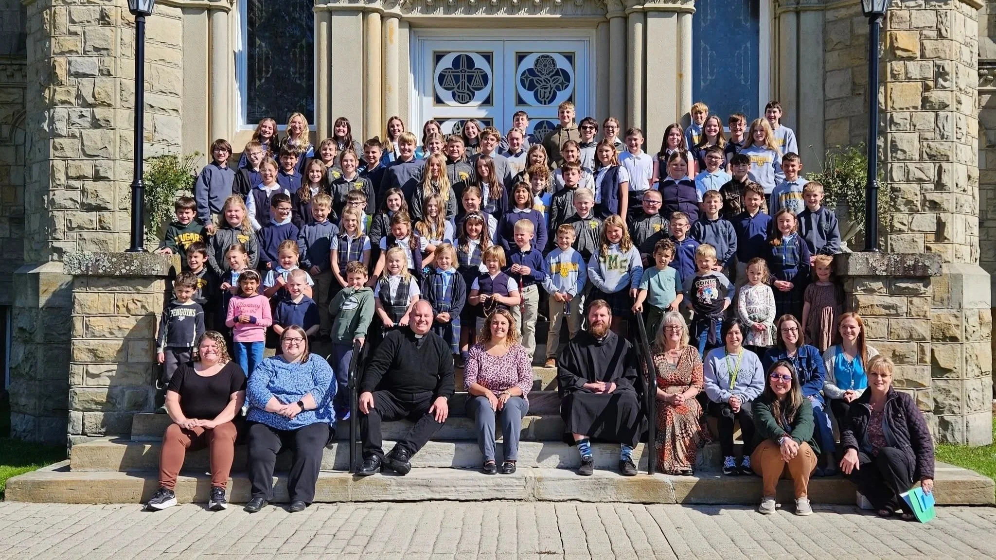 Group photo of students, teachers, and clergy members on the steps outside a stone building with decorative glass doors, taken on a sunny day.
