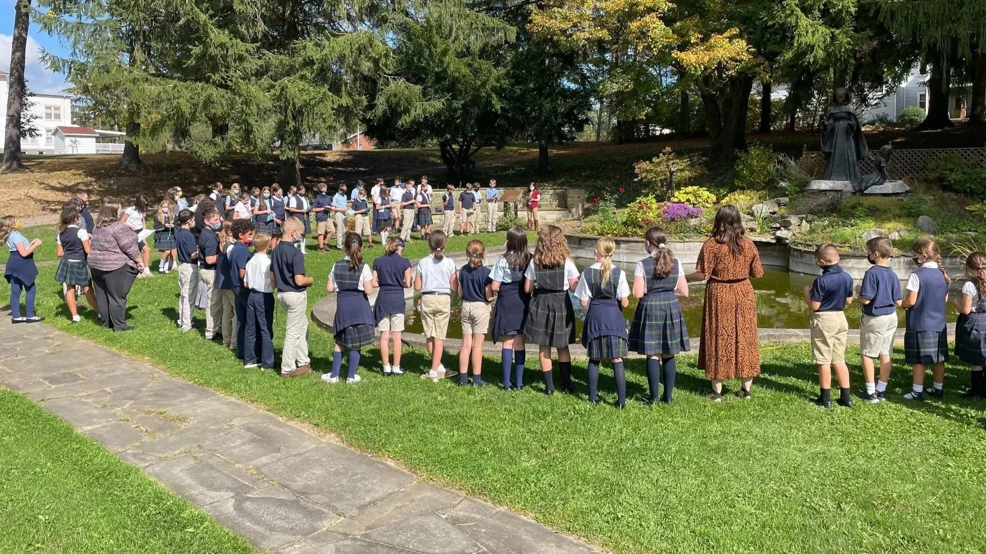 A group of school children and a teacher gather outside near a statue on a sunny day, surrounded by trees and greenery.