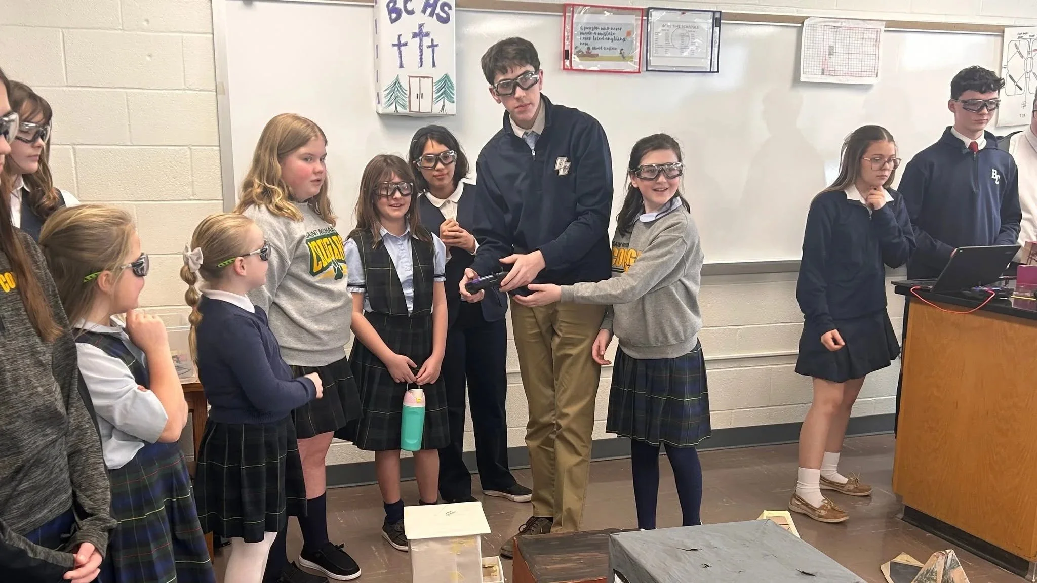 A group of elementary school students in plaid skirts and uniforms gather around a male teacher wearing safety goggles, who is demonstrating a science experiment in a classroom. The students watch attentively, some with curiosity and interest.