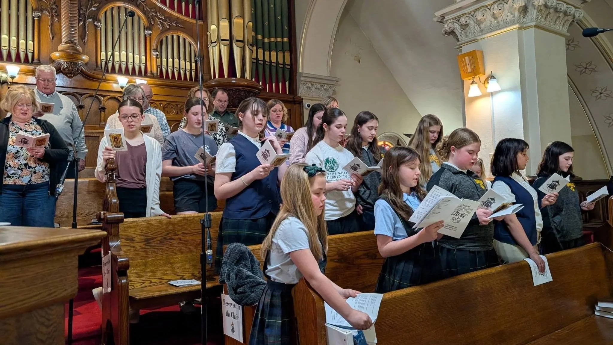 A group of young girls and a few adults standing and singing in a church during a choir practice or performance, holding sheet music, with ornate wood and decorative arches in the background.