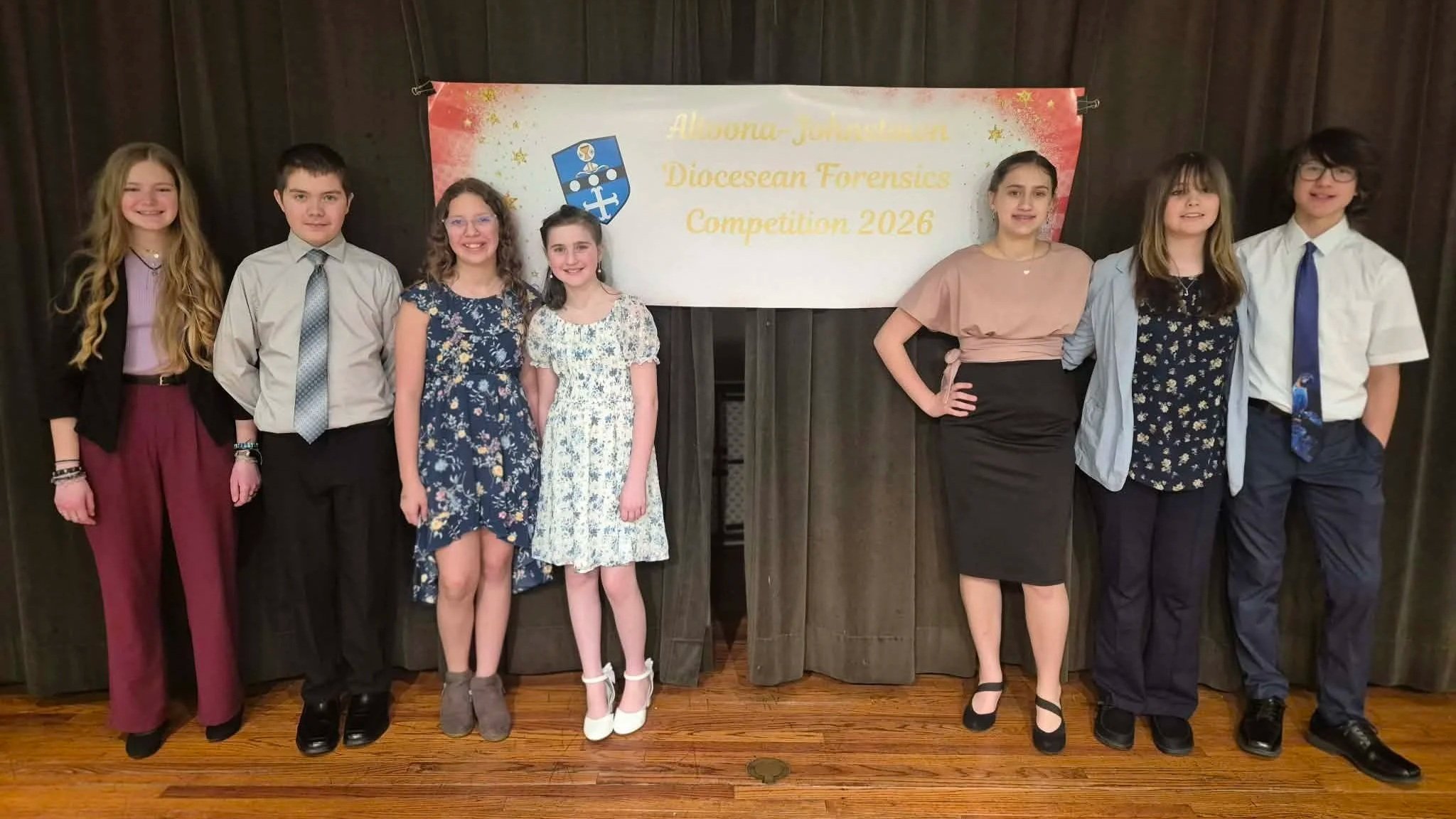 A group of seven young people, five girls and two boys, standing in front of a banner that reads 'Aloha-January Diocesan Forensics Competition 2026.' They are dressed in semi-formal attire and smiling, standing on a wooden floor.