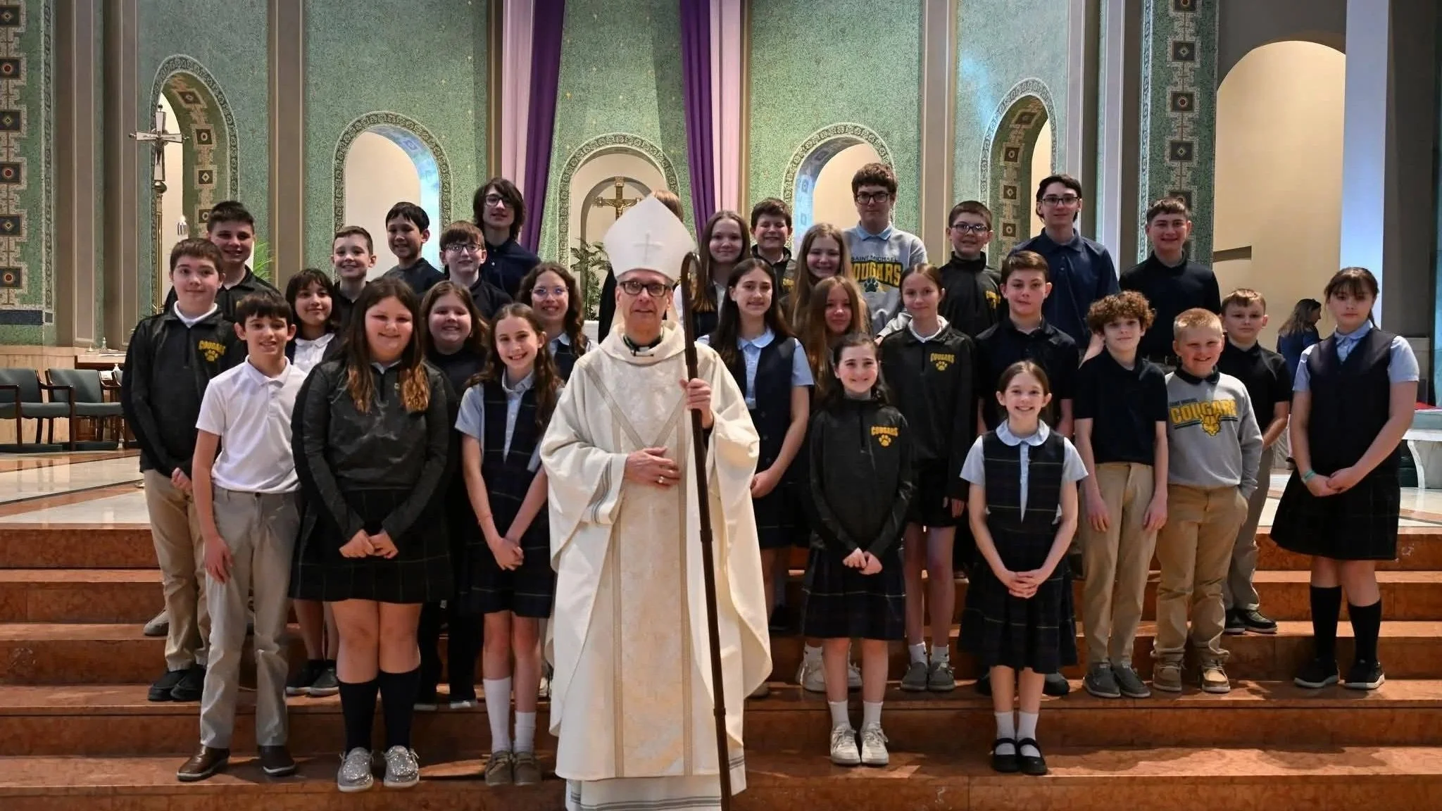 Group of children with a bishop inside a church with green patterned walls and a crucifix in the background.
