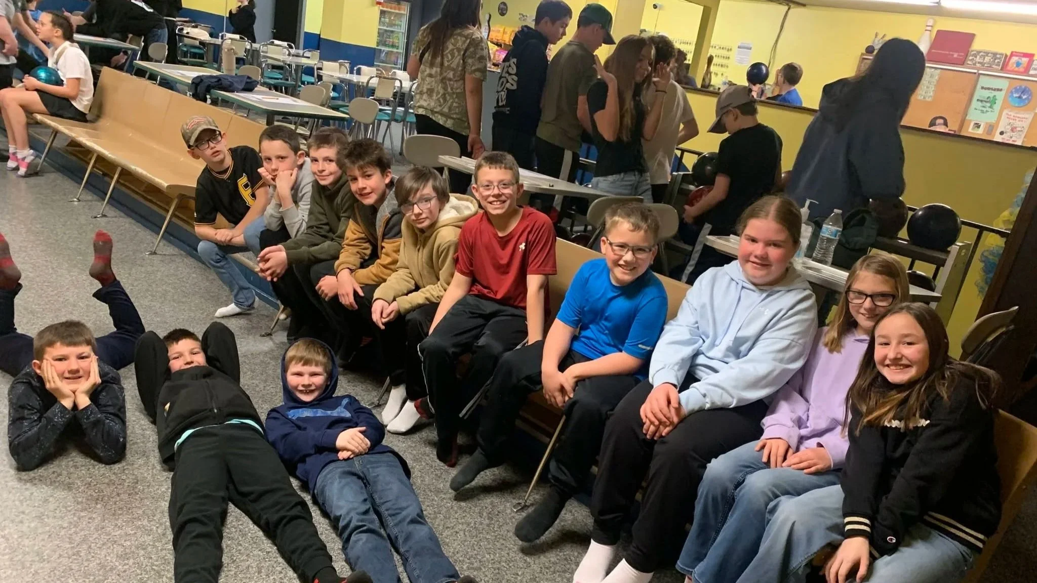 A group of children sitting and lying on the floor and chairs at a bowling alley, smiling for the camera. Some children are holding bowling balls, and others are looking around or talking.