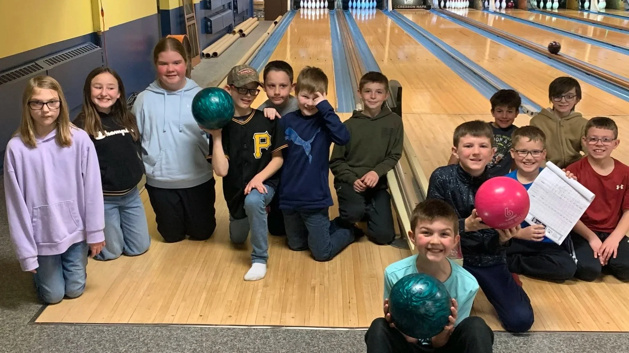 Group of children at a bowling alley, some holding bowling balls, smiling, with bowling lanes in the background.