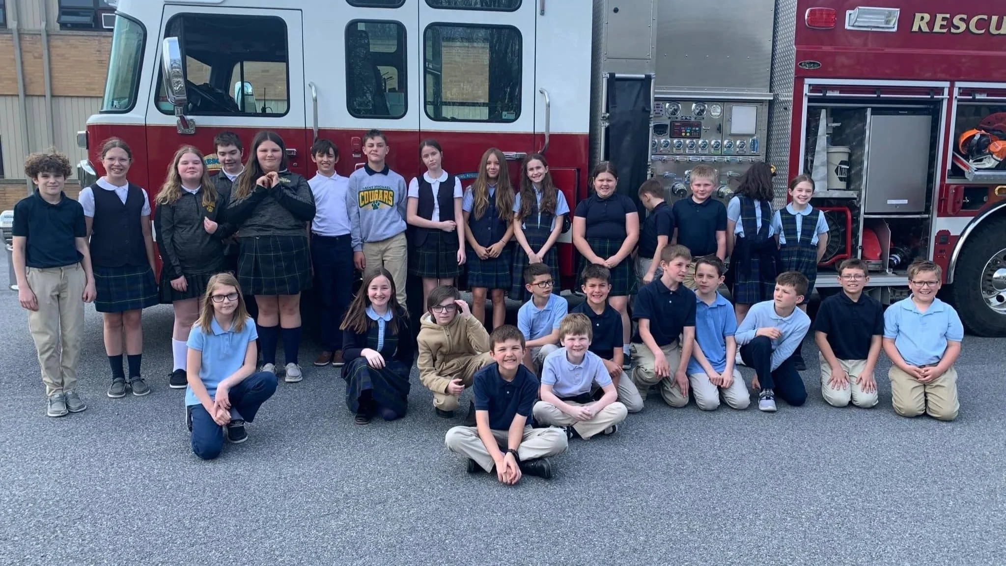 A group of children and a few adults preschool age, standing and sitting in front of a fire truck for a photo. The children are wearing school uniforms or casual clothing. The fire truck is red and white, equipped with various lights, gauges, and equ