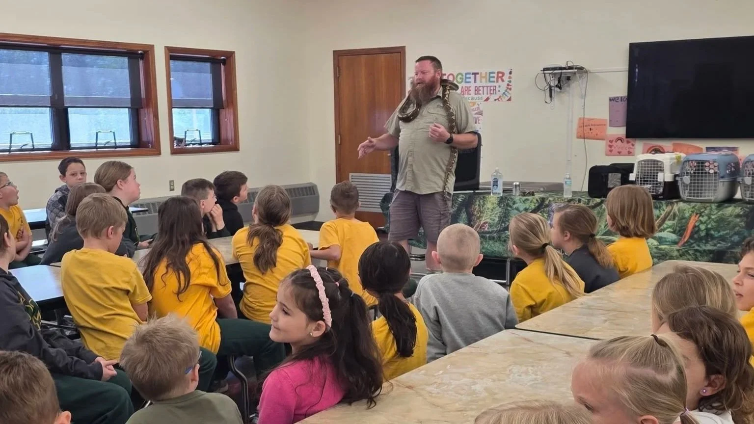 A man with a snake around his shoulders speaking to children in a classroom.