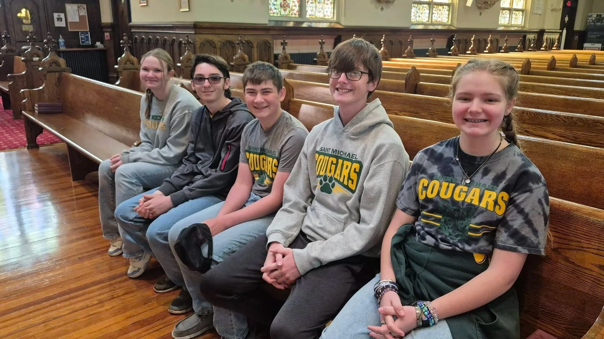 Five children sitting on a wooden church pew inside a church, wearing gray shirts with 'Saint Michael Cougars' logo, smiling at the camera.