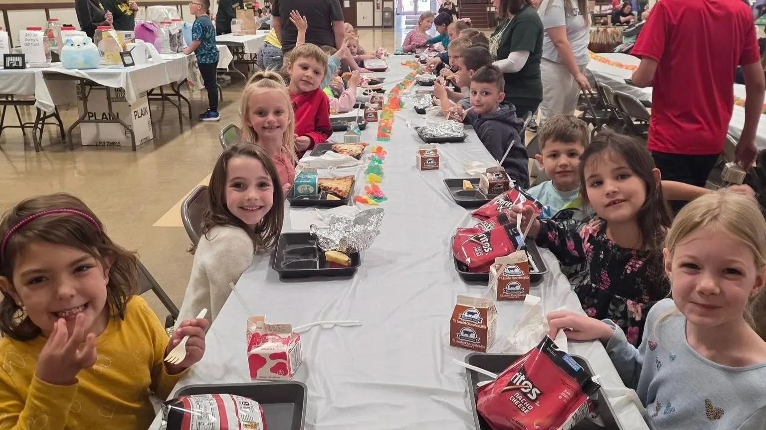 Children sitting at a long table enjoying a meal with pizza, snacks, and drinks, likely celebrating a party or event.