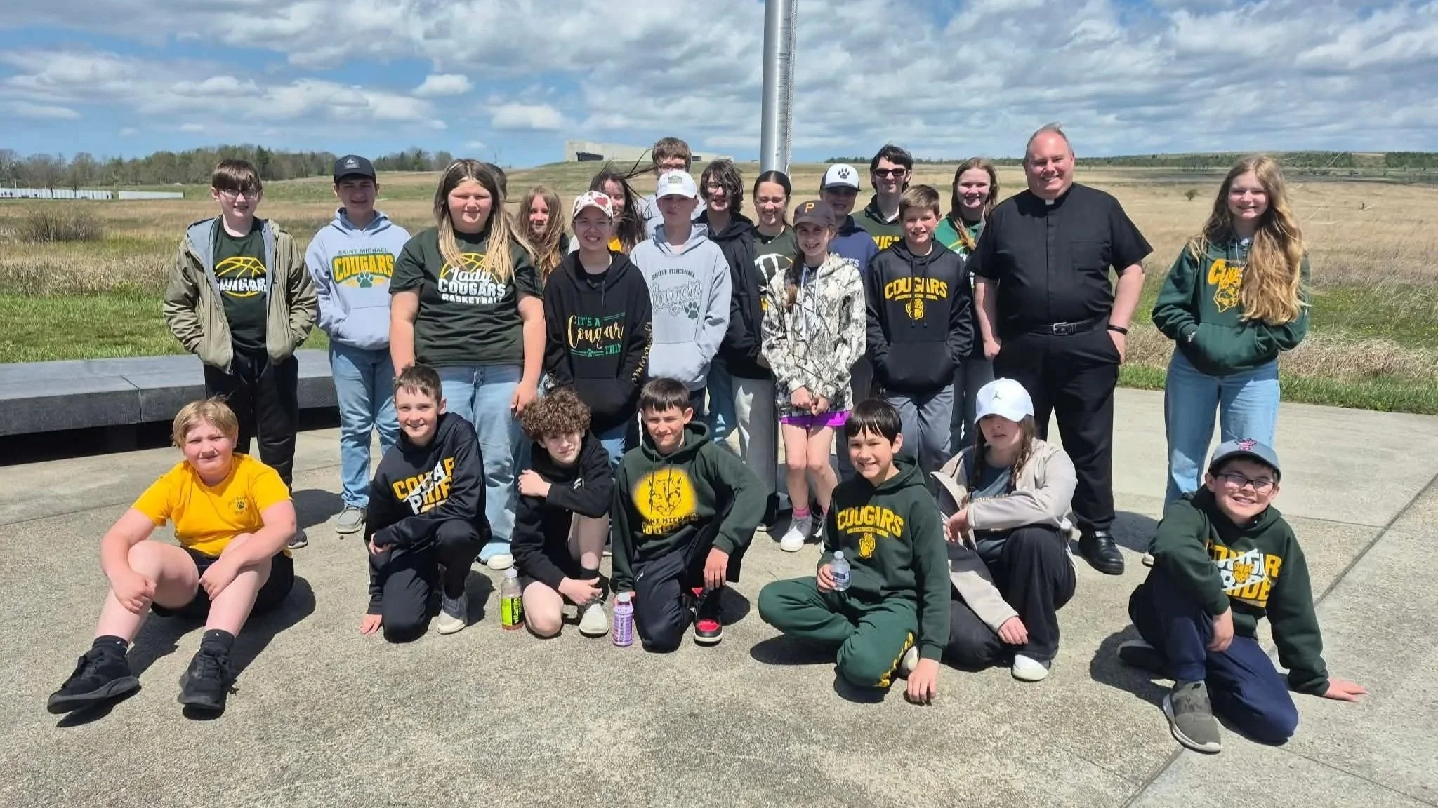 A large group of children and adults posing outdoors on a cloudy day, many wearing 'Cougars' or school-themed apparel, with a grassy field and some structures in the background.