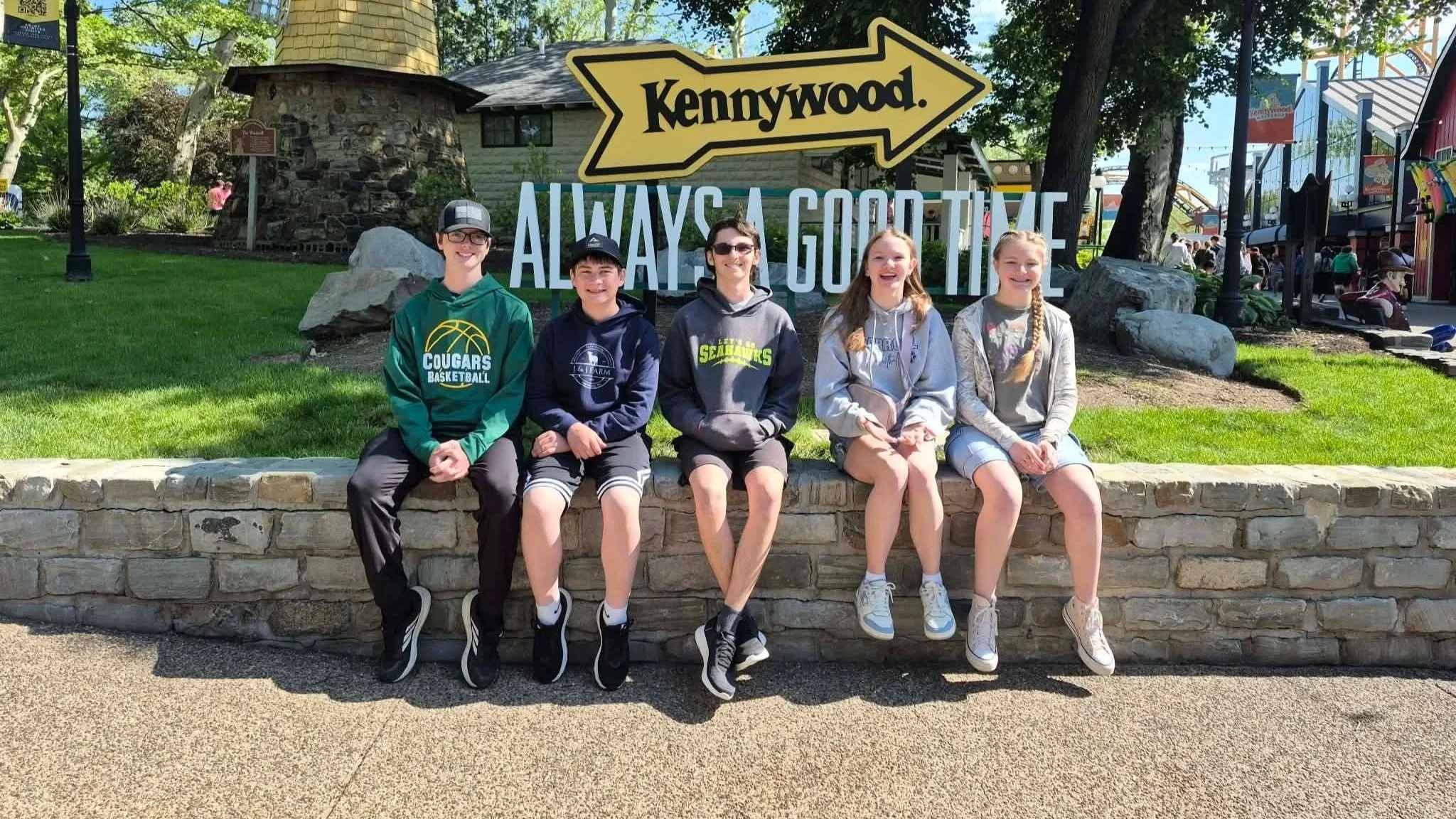 Five children sitting on a stone ledge in front of a sign that says 'Kennywood' with an arrow and 'Always a good time' in the background at Kennywood amusement park.