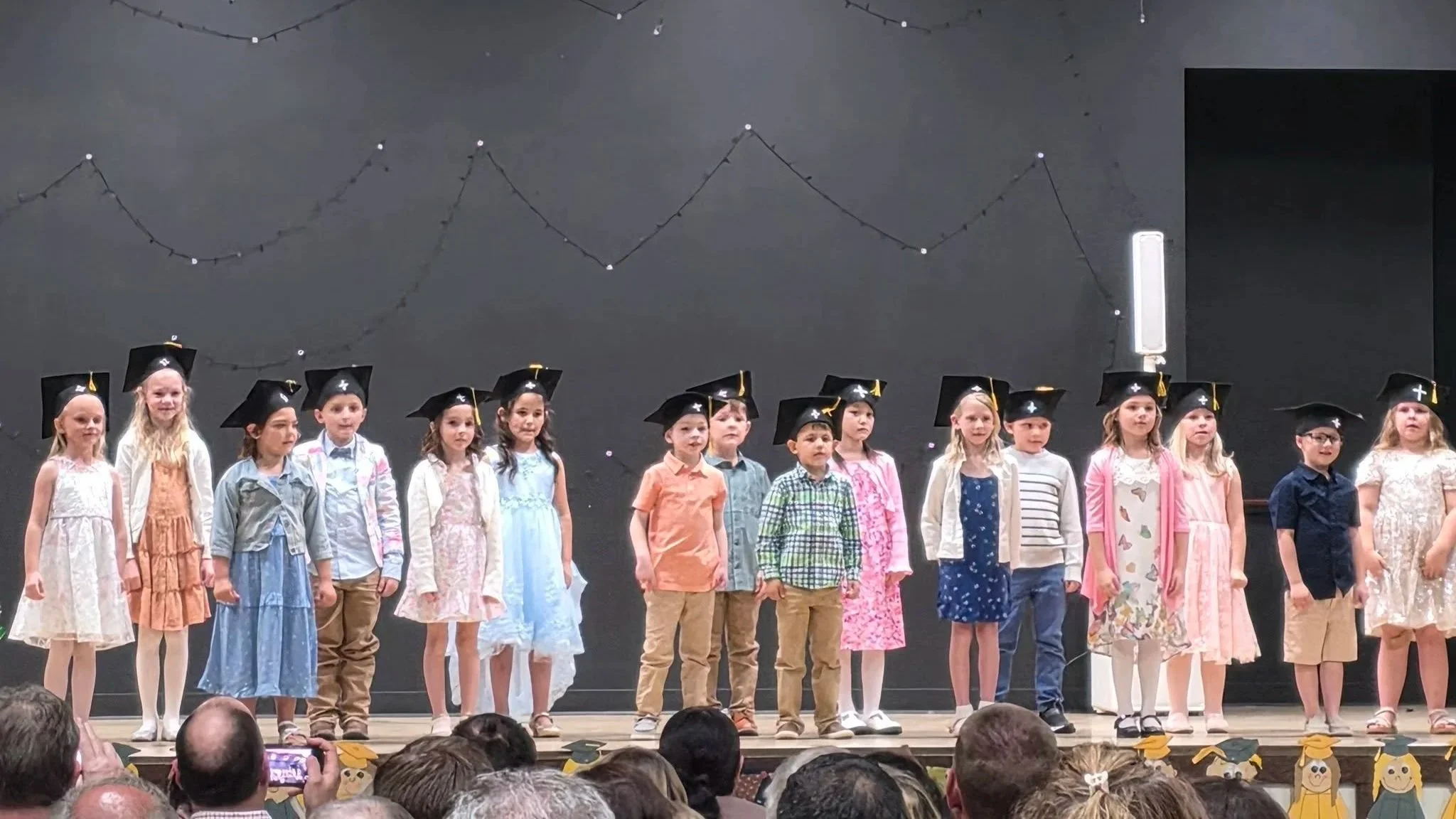 A group of children dressed in colorful clothing and graduation caps standing on stage during a graduation or school event, with an audience in front.