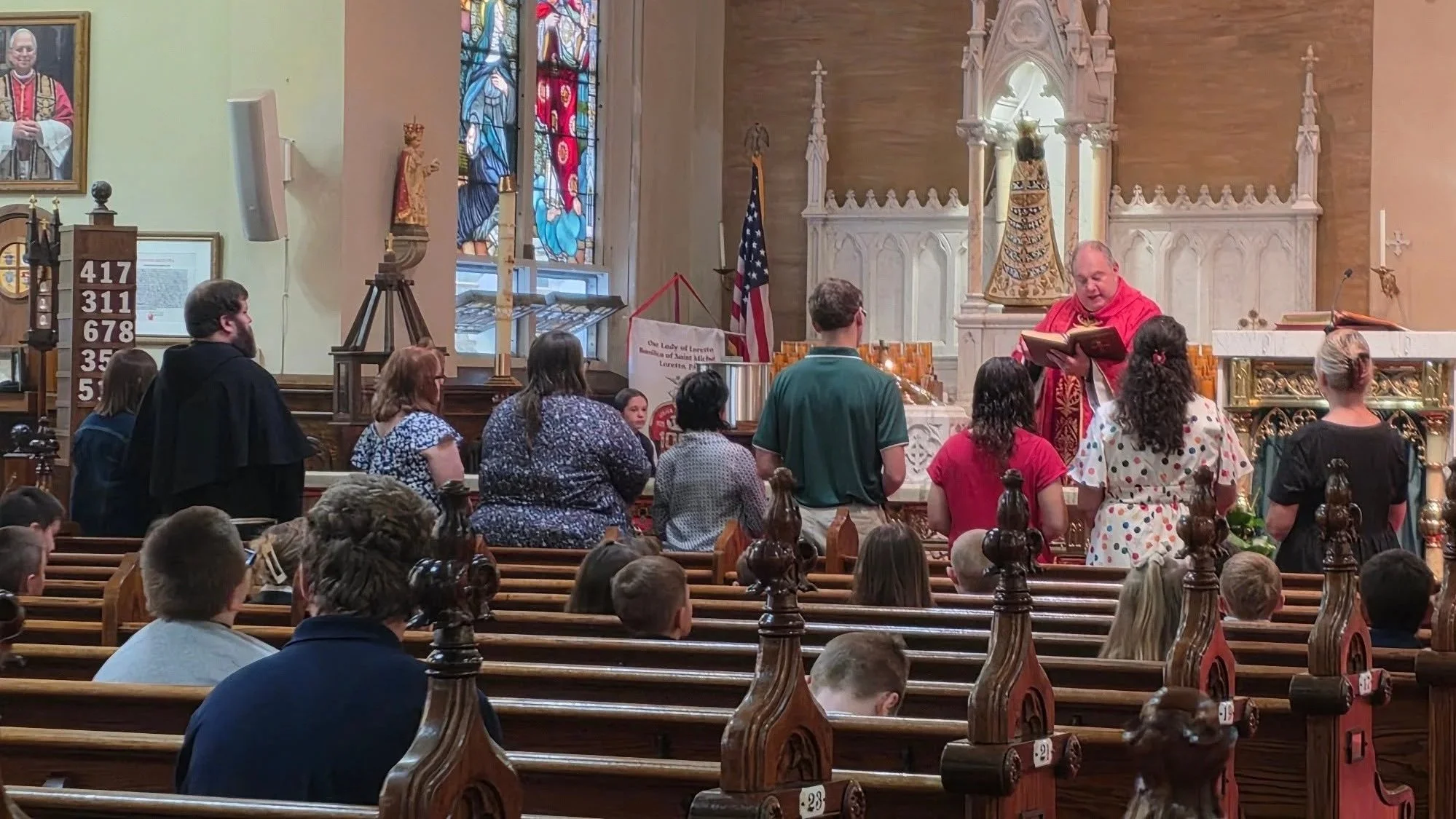 Inside a church with stained glass windows and intricate woodwork, a priest in red robes is reading from a book to a congregation of diverse people, some standing and some seated in wooden pews.
