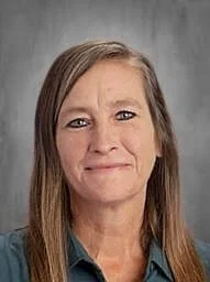 A woman with shoulder-length light brown hair and a slight smile, wearing a teal shirt, standing in front of a gray background.