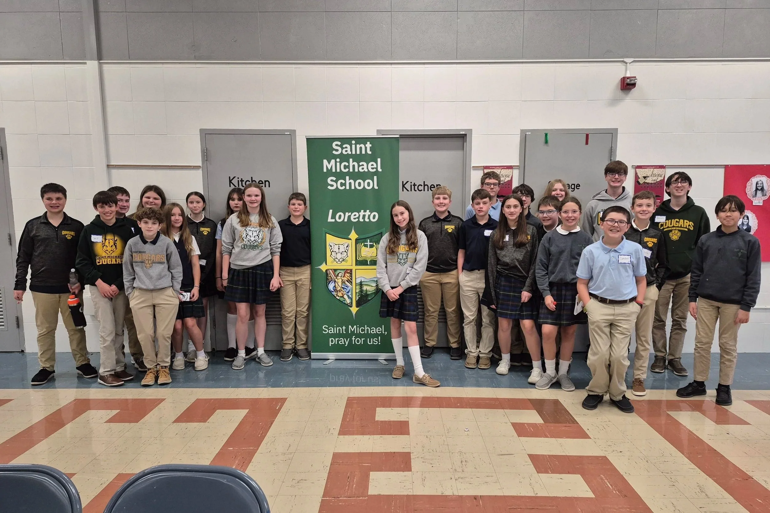 Group of students standing indoors in front of a green school banner that reads 'Saint Michael School' and 'Loretto.' The students are wearing school uniforms, with some wearing black or gray sweaters with the school logo, and others in collared shirts and khakis. The students are smiling and standing in a line, with the school hallway and closed doors visible in the background.